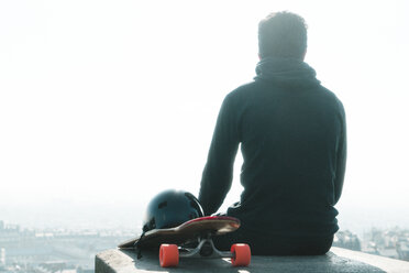 Rear view of man with skateboard sitting on column against clear sky - CAVF57062