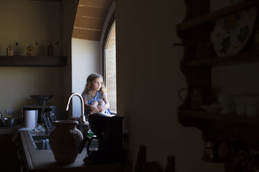 Mädchen, das durch das Fenster schaut, während es in der Küche zu Hause sitzt - CAVF57077