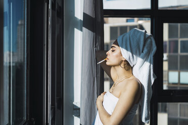Side view of young woman smoking cigarette while being wrapped in towel at home - CAVF57125