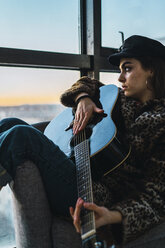 Nachdenkliche junge Frau spielt Gitarre, während sie zu Hause auf dem Sofa vor dem Fenster sitzt - CAVF57128