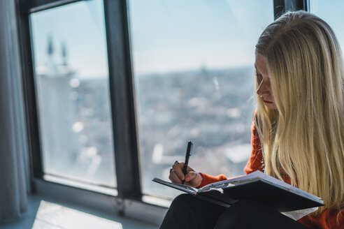Junge Frau beim Skizzieren auf einem Buch, während sie zu Hause am Fenster sitzt - CAVF57132