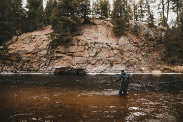 Rear view of male hiker with backpack fishing in river by mountain - CAVF57143