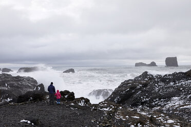 Rückansicht eines Vaters mit Tochter, der auf einem Felsen bei Dyrholaey Island steht und auf das Meer blickt, bei bewölktem Himmel - CAVF57156
