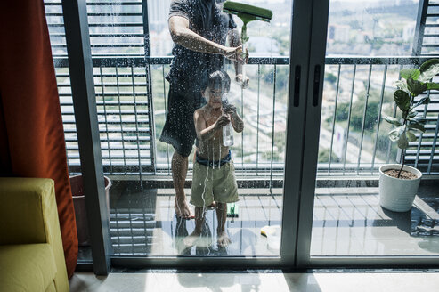 Tiefschnitt von Vater mit Sohn beim Fensterputzen auf dem Balkon durch Glas gesehen - CAVF57160