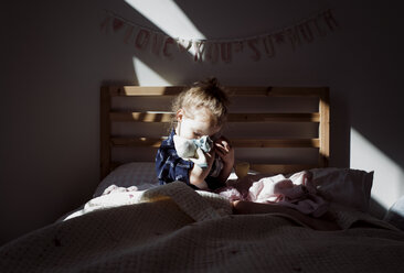Mädchen umarmt Puppe auf dem Bett sitzend in der Dunkelkammer zu Hause - CAVF57182