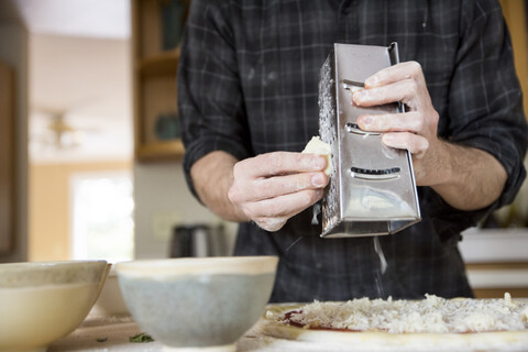 Mittelteil eines Mannes, der auf einer Kücheninsel zu Hause Käse über eine Pizza reibt, lizenzfreies Stockfoto