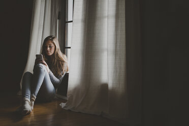 Teenager-Mädchen in voller Länge bei der Benutzung eines Smartphones, während sie zu Hause am Fenster sitzt - CAVF57235