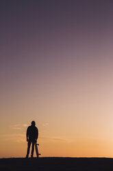 Silhouette Mann mit Skateboard steht gegen dramatischen Himmel bei Sonnenuntergang - CAVF57244