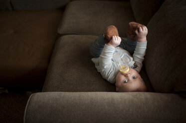 Hochformatiges Porträt eines kleinen Jungen, der Milch aus der Flasche trinkt, während er zu Hause auf dem Sofa liegt - CAVF57287