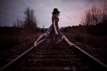Seitenansicht einer Frau, die auf Eisenbahnschienen vor einem bewölkten Himmel bei Sonnenuntergang steht - CAVF57310