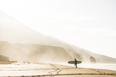 Man with surfboard standing on shore at beach against clear sky during foggy weather - CAVF57364