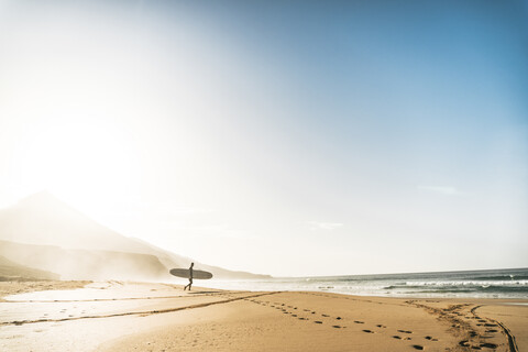 Mann mit Surfbrett geht am Strand gegen den Himmel bei nebligem Wetter zum Meer, lizenzfreies Stockfoto