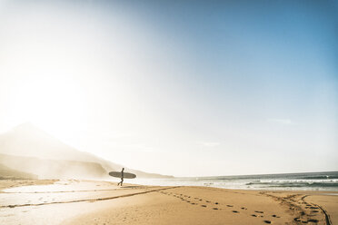 Man with surfboard walking towards sea at beach against sky during foggy weather - CAVF57365