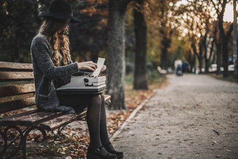Seitenansicht einer Schriftstellerin, die auf einer Parkbank sitzend eine Schreibmaschine benutzt, lizenzfreies Stockfoto