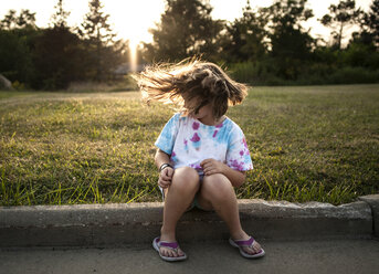 Verspieltes Mädchen, das sich die Haare zuwirft, während es im Park bei Sonnenuntergang sitzt - CAVF57411