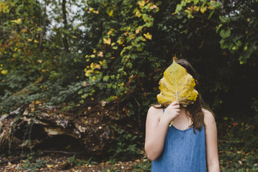 Mädchen hält sich ein Blatt vor das Gesicht, während sie im Herbst im Park steht - CAVF57441