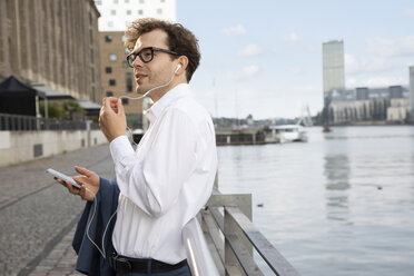 Germany, Berlin, businessman using smartphone and earphones outdoors - FKF03103