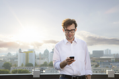 Germany, Berlin, smiling businessman on roof terrace looking at smartphone in the evening stock photo