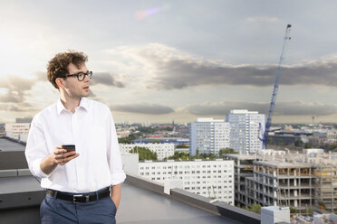 Deutschland, Berlin, Geschäftsmann mit Handy auf Dachterrasse stehend mit Blick auf Aussicht - FKF03127