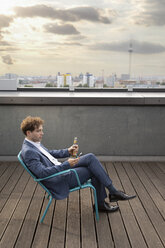 Deutschland, Berlin, Geschäftsmann entspannt mit Getränk auf Dachterrasse bei Sonnenuntergang und schaut auf sein Handy - FKF03133