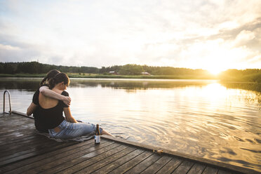 Female friends embracing while sitting on jetty over lake against sky during sunset - MASF09702