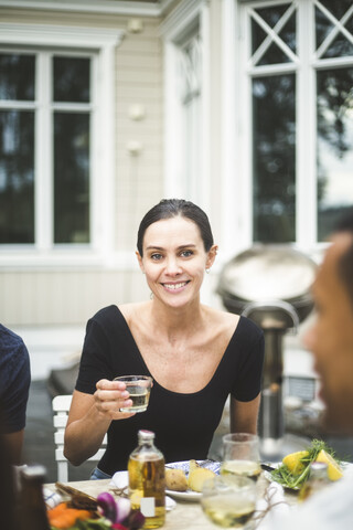 Porträt einer lächelnden erwachsenen Frau, die ein Getränk in einem Glas hält, während sie am Esstisch vor einer Villa sitzt, lizenzfreies Stockfoto