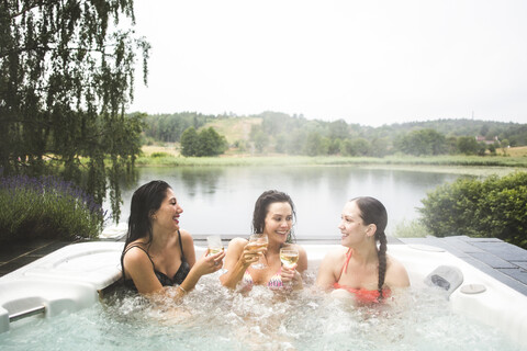 Fröhliche Freundinnen genießen Wein im Whirlpool am See während eines Wochenendausflugs, lizenzfreies Stockfoto