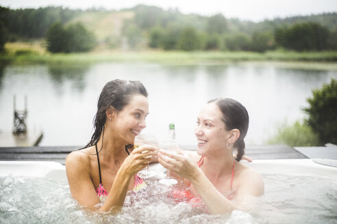 Lächelnde Freundinnen stoßen mit Weingläsern an, während sie sich im Whirlpool am See vergnügen - MASF09725
