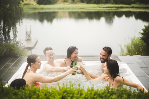 Cheerful male and female friends toasting drinks in hot tub against lake during weekend getaway stock photo