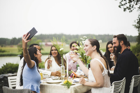 Fröhliche junge Frau macht ein Selfie mit Freunden während einer Dinnerparty im Hinterhof, lizenzfreies Stockfoto