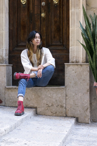 Portrait of young woman wearing red boots sitting on step in front of entry door stock photo