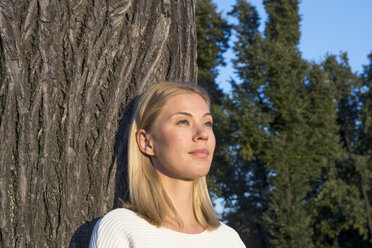 Porträt einer träumenden blonden Frau, die sich an einen Baumstamm lehnt und in die Ferne schaut - LMJF00032