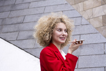 Porträt einer lächelnden blonden Frau mit Locken, die einen roten Anzug trägt und mit einem Mobiltelefon spricht - LMJF00050