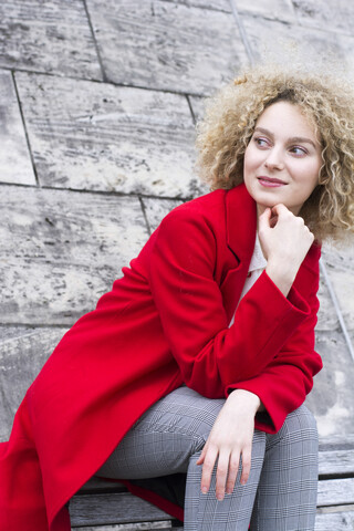 Porträt einer lächelnden blonden Frau mit Locken und rotem Mantel, die etwas beobachtet, lizenzfreies Stockfoto