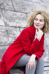 Porträt einer lächelnden blonden Frau mit Locken und rotem Mantel, die etwas beobachtet - LMJF00052