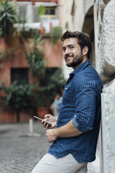 Portrait of smiling man with smartphone leaning against facade looking at distance - BOYF01075