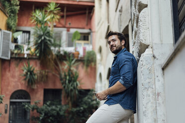 Bearded man with smartphone leaning against facade looking at distance - BOYF01077