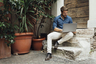 Smiling businessman sitting on steps in a courtyard using laptop - BOYF01092