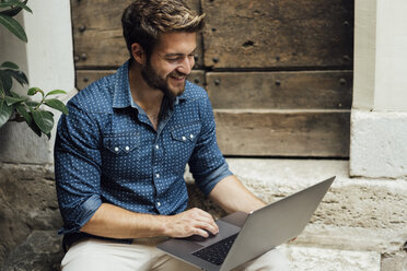 Smiling businessman sitting on steps in a courtyard using laptop - BOYF01093