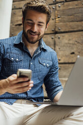 Portrait of smiling businessman with laptop looking at smartphone outdoors - BOYF01095