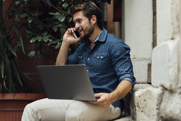 Smiling businessman on the phone sitting on steps in a courtyard with laptop - BOYF01098