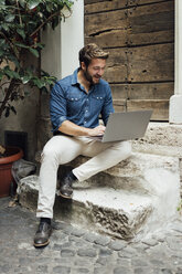 Lachender Geschäftsmann, der auf einer Treppe in einem Innenhof sitzt und einen Laptop benutzt - BOYF01100