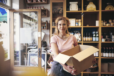 Porträt einer selbstbewussten Verkäuferin, die eine Pappschachtel gegen ein Regal in einem Feinkostladen trägt - MASF09774