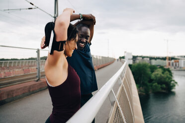 Smiling male and female athletes stretching hands on footbridge over sea against sky - MASF09885