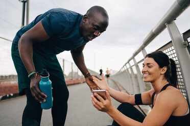 Female athlete smiling while showing mobile phone to sportsman on footbridge - MASF09886