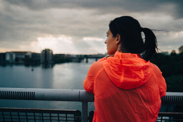 Rückansicht einer Sportlerin im Regenmantel mit Blick auf die Stadt, während sie bei Sonnenuntergang auf einer Fußgängerbrücke über dem Meer steht - MASF09894