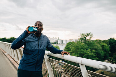 Portrait of thirsty sportsman drinking from bottle while standing on footbridge against sky - MASF09899