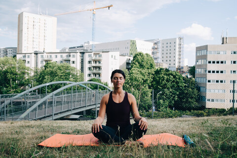 Junge Frau meditiert auf einem Feld vor einem Gebäude in der Stadt, lizenzfreies Stockfoto