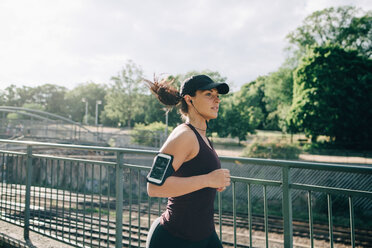 Confident sportswoman listening music through in-ear headphones while jogging on bridge in city - MASF09914