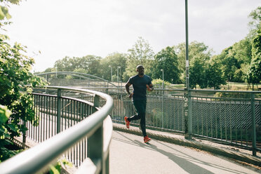 Full length of mid adult sportsman jogging on footbridge in city during sunny day - MASF09916